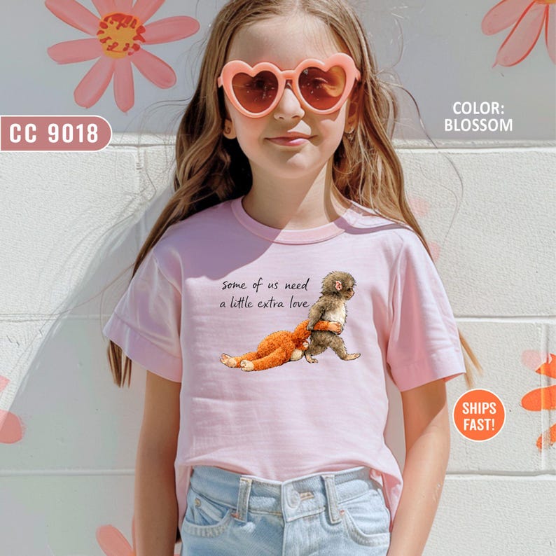 May include: A pale pink t-shirt featuring the phrase "some of us need a little extra love" and a graphic of a monkey hugging a stuffed toy. The shirt also has the text "SHIPS FAST!" and "COLOR: BLOSSOM".