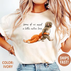 May include: Ivory t-shirt with a cartoon illustration of two monkeys, one carrying the other. The text "some of us need a little extra love" is above the image. The shirt is ivory in colour.