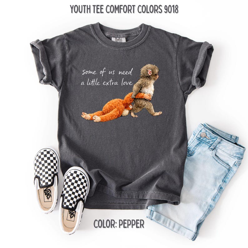 May include: A dark grey youth tee featuring the text "some of us need a little extra love" and an illustration of a monkey hugging a teddy bear. The image includes light blue denim shorts and black and white checkered slip-on shoes.