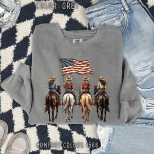 May include: A grey Comfort Colors sweatshirt featuring an illustration of four cowgirls riding horses, with one holding an American flag. The text "COLOR: GREY" and "COMFORT COLORS 1566" are printed on the sweatshirt.
