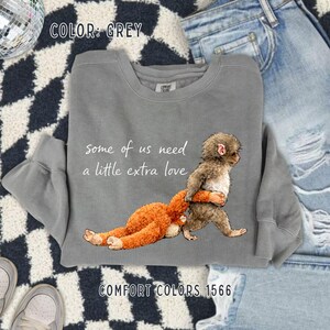 May include: Grey sweatshirt featuring the text "some of us need a little extra love" and a graphic of two monkeys. One monkey is brown and the other is orange. The sweatshirt is Comfort Colors 1566.