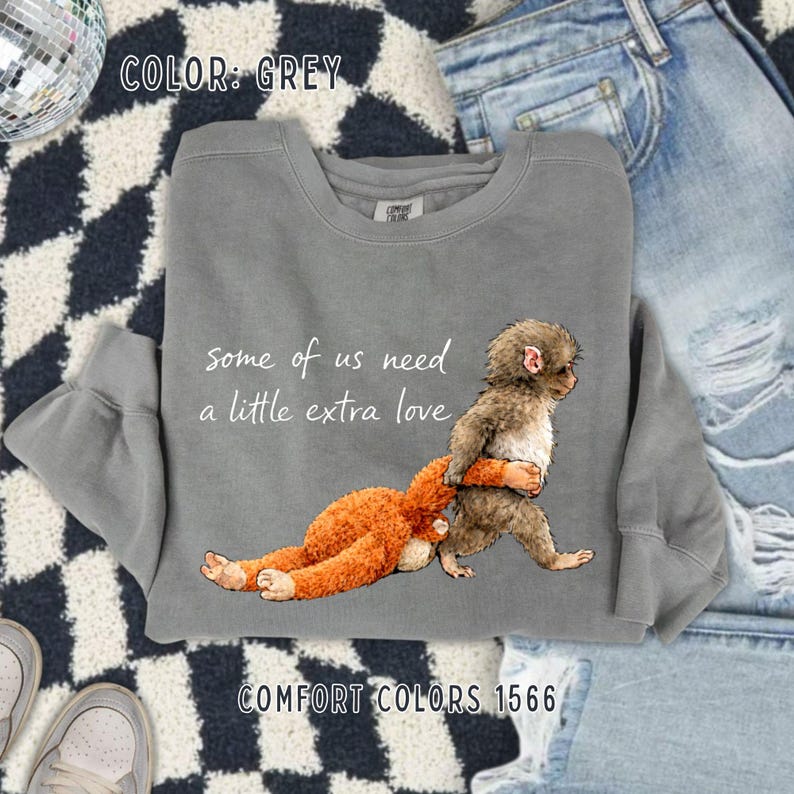 May include: Grey sweatshirt featuring the text "some of us need a little extra love" and a graphic of two monkeys. One monkey is brown and the other is orange. The sweatshirt is Comfort Colors 1566.