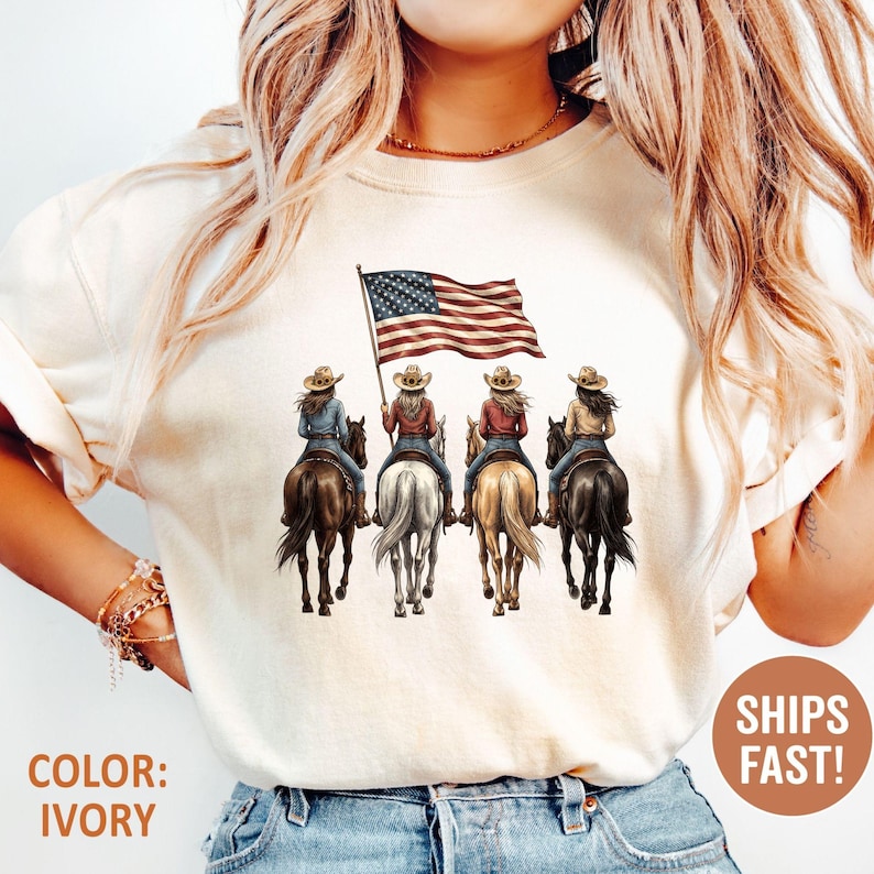 May include: Ivory-colored t-shirt featuring a graphic of four cowgirls riding horses, with an American flag. The shirt has the text "COLOR: IVORY" and a circular graphic that says "SHIPS FAST!"