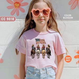 May include: A light pink t-shirt featuring a graphic of four cowgirls riding horses, holding an American flag. The text "CC 9018" is on the left, and "COLOR: BLOSSOM" is on the right. The shirt is worn by a person wearing heart-shaped sunglasses.