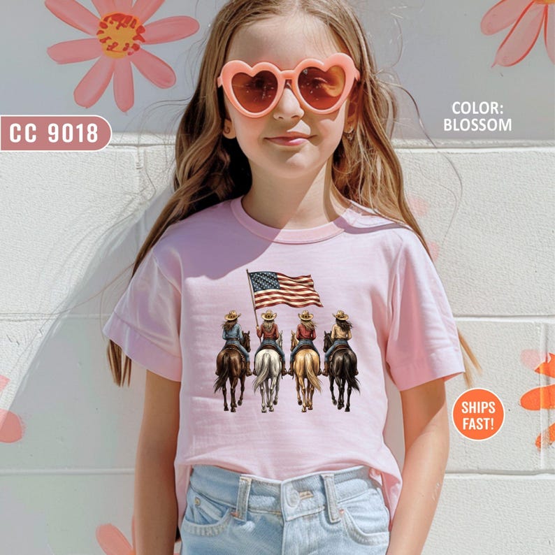 May include: A light pink t-shirt featuring a graphic of four cowgirls riding horses, holding an American flag. The text "CC 9018" is on the left, and "COLOR: BLOSSOM" is on the right. The shirt is worn by a person wearing heart-shaped sunglasses.