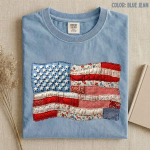 May include: Blue jean colored t-shirt with a sewn-on American flag design. The flag features white stars on a blue field and red and white stripes made of various patterned fabrics. The shirt has a crew neck and a Comfort Colors tag.