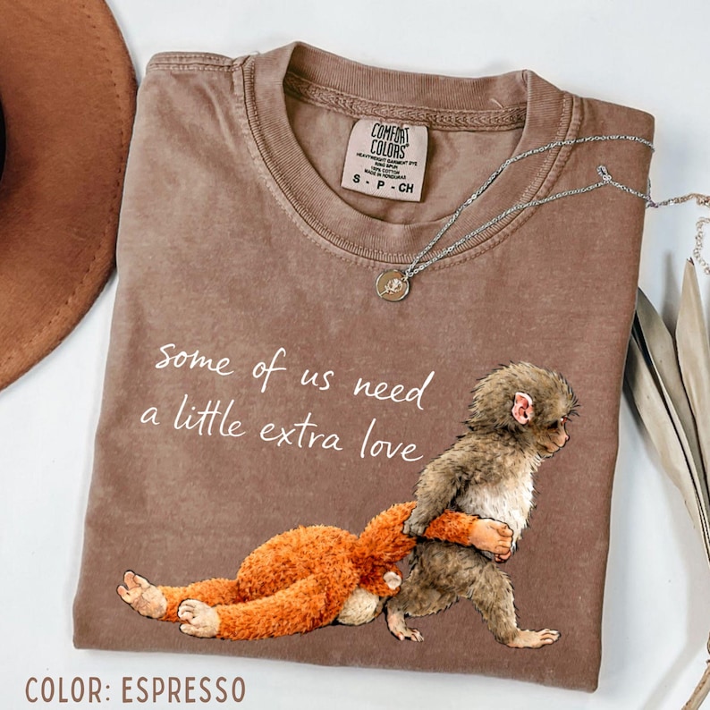 May include: Espresso-coloured t-shirt featuring the text "some of us need a little extra love" and a graphic of a monkey hugging a stuffed toy. The shirt has a crew neck and short sleeves. A silver necklace with a pendant is also visible.