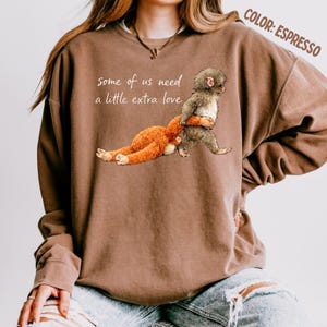 May include: Espresso-coloured sweatshirt featuring a graphic of a monkey hugging a stuffed animal. The text "some of us need a little extra love" is printed on the front. The sweatshirt has long sleeves and a crew neck.