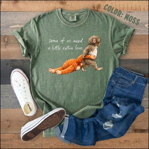 May include: A moss green t-shirt featuring the text "some of us need a little extra love" and a graphic of a monkey hugging a stuffed toy. The image also shows blue jeans and white trainers.