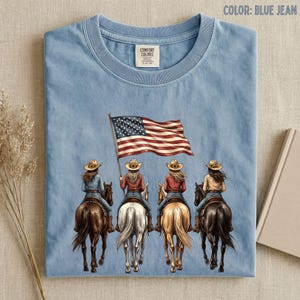 May include: Blue jean colored t-shirt featuring an illustration of four cowgirls riding horses, with one holding an American flag. The shirt has a crew neck and a small tag on the upper left. The words "COLOR: BLUE JEAN" are printed at the top.