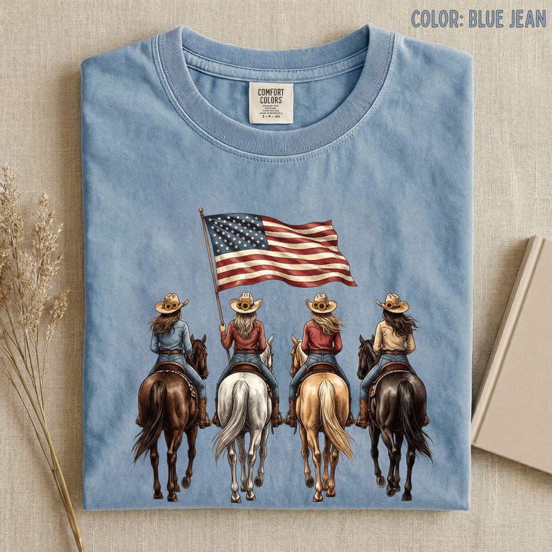 May include: Blue jean colored t-shirt featuring an illustration of four cowgirls riding horses, with one holding an American flag. The shirt has a crew neck and a small tag on the upper left. The words "COLOR: BLUE JEAN" are printed at the top.