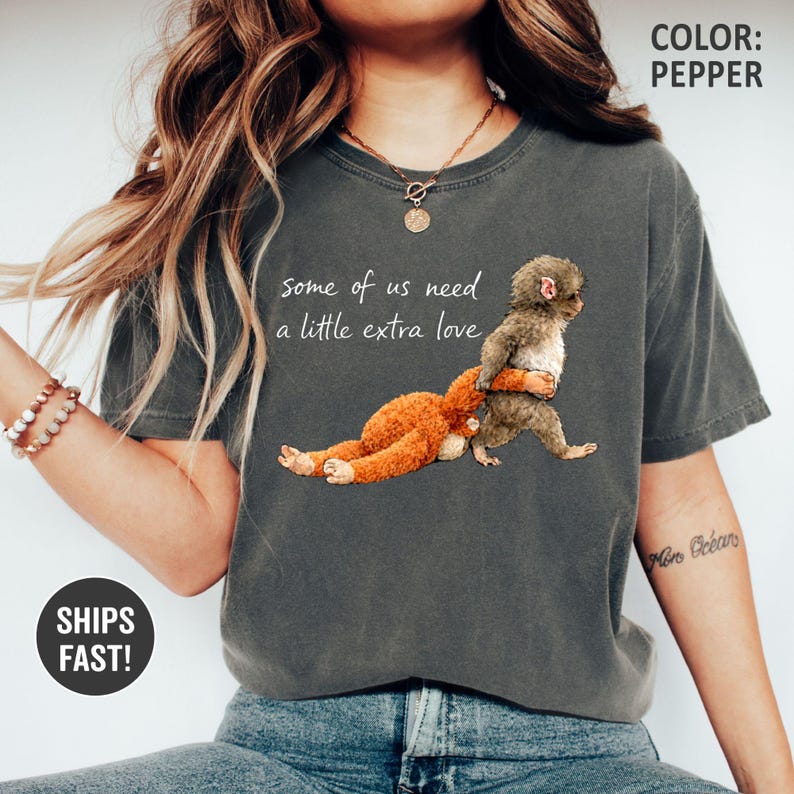 May include: A dark grey t-shirt with the text "some of us need a little extra love" and a graphic of a brown toy hugging a grey animal. The shirt is a casual style and the colour is pepper.