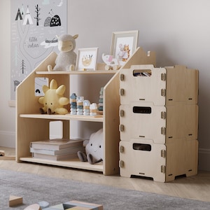 Può includere: Una libreria in legno chiaro a tre ripiani, con peluche, quadri incorniciati e libri. A destra c'è una pila di tre contenitori portaoggetti coordinati. La libreria ha un design a gradini unico, perfetto per la cameretta.