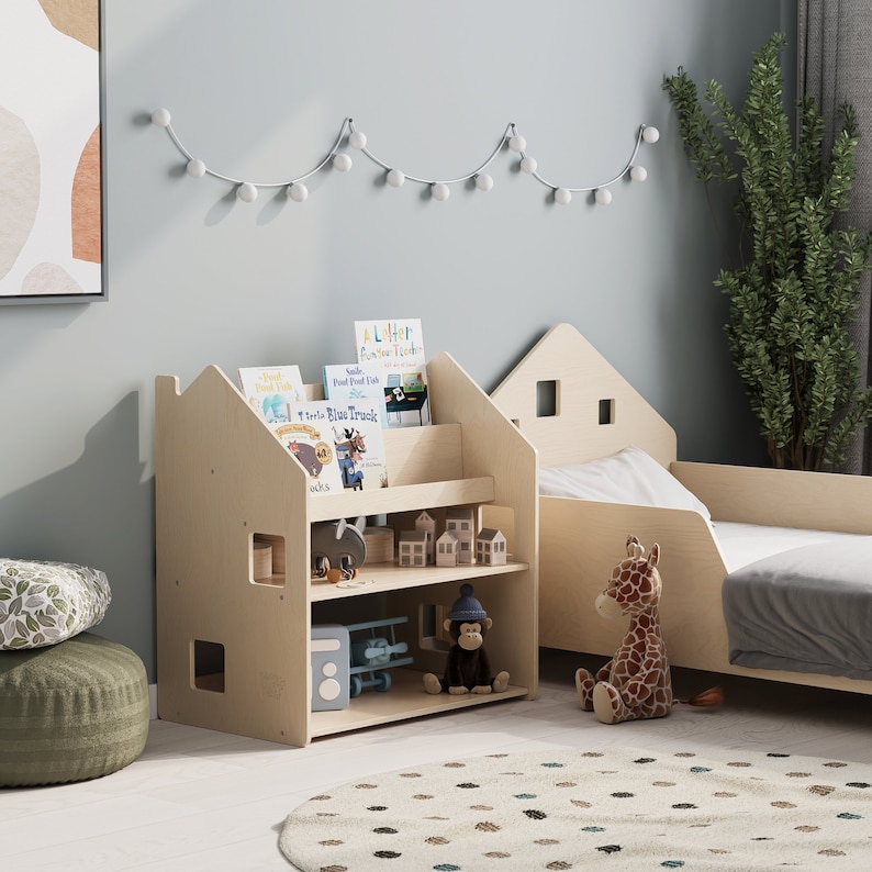 May include: A light wood children's bookshelf with three shelves, featuring a house-shaped design. The bookshelf is filled with children's books and toys, including a toy airplane, a stuffed monkey, and a stuffed giraffe. The bookshelf is in a room with a light blue wall and a white string of lights.