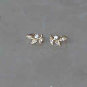 May include: A pair of gold-colored leaf-shaped earrings with small, clear gemstones. The earrings are displayed on a gray surface. The brand name "Winnies & ATOWN - jewelry" is visible in the lower center.