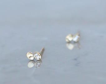 Puede incluir: Un par de pendientes de botón dorados, cada uno con un pequeño diseño en forma de hoja y una gema transparente. Los pendientes se muestran sobre una superficie gris claro. La marca "Winnies ATOWN JEWELRY" es visible.