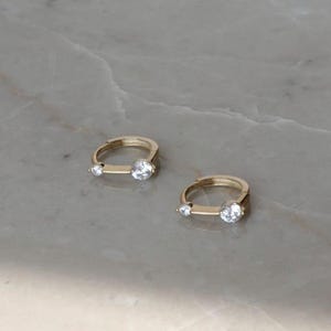 Puede incluir: Dos pendientes de aro dorados, cada uno con una peque&ntilde;a gema transparente y una gema transparente y redonda m&aacute;s grande. Los pendientes se muestran sobre una superficie clara, similar al m&aacute;rmol. La marca "Winnies & ATOWN JEWELRY" es visible en el fondo.