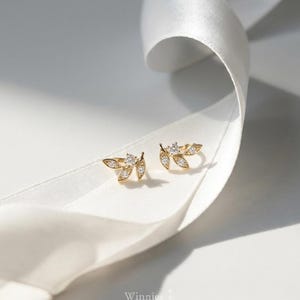 May include: A pair of gold-colored leaf-shaped stud earrings with small, clear gemstones. The earrings are displayed on a white ribbon against a white background. The brand name "Winnies ATown" is visible.