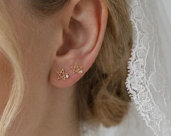 Puede incluir: Pendientes de botón dorados en forma de estrella, cada uno con un pequeño acento de diamante. Los pendientes se llevan en la oreja de una persona, con un velo blanco visible en el fondo. Joyería delicada.