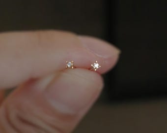 Puede incluir: Un par de pequeños pendientes de botón dorados en forma de estrella, cada uno con una pequeña gema transparente en el centro. Los pendientes se sostienen entre los dedos sobre un fondo oscuro. La marca "Winnies & Atown Jewelry" es visible.