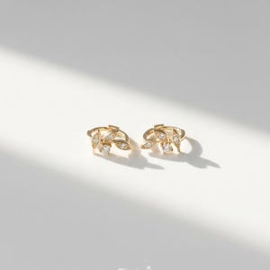 May include: A pair of gold-colored leaf-shaped earrings with small, clear gemstones. The earrings are on a white surface, with a shadow cast across them. The earrings have a delicate, elegant design.