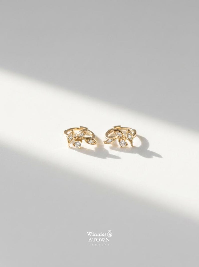 May include: A pair of gold-colored leaf-shaped earrings with small, clear gemstones. The earrings are on a white surface, with a shadow cast across them. The earrings have a delicate, elegant design.