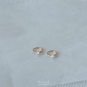 Puede incluir: Dos pendientes de aro dorados, cada uno con una sola gema transparente. Los pendientes se muestran sobre una superficie de color claro. La marca "Winnies & A Town" es visible.
