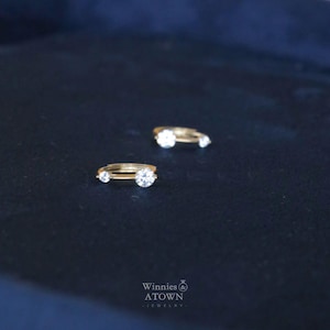 Puede incluir: Dos anillos de oro con piedras preciosas redondas y transparentes se muestran sobre una superficie azul oscuro. Un anillo tiene una piedra central m&aacute;s grande flanqueada por piedras m&aacute;s peque&ntilde;as. Los anillos son de Winnies & ATOWN Jewelry.