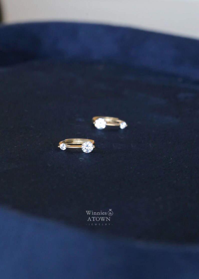 Puede incluir: Dos anillos de oro con piedras preciosas redondas y transparentes se muestran sobre una superficie azul oscuro. Un anillo tiene una piedra central m&aacute;s grande flanqueada por piedras m&aacute;s peque&ntilde;as. Los anillos son de Winnies & ATOWN Jewelry.