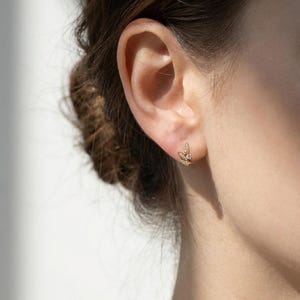 May include: A close-up of a gold-colored stud earring with a leaf design, set with small, clear gemstones. The earring is worn on an ear, with the brand name "Winnies & A Town Jewelry" visible in the lower right corner.
