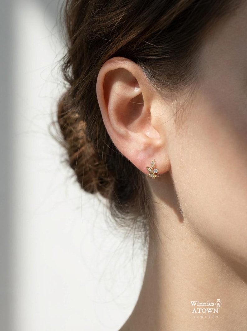 May include: A close-up of a gold-colored stud earring with a leaf design, set with small, clear gemstones. The earring is worn on an ear, with the brand name "Winnies & A Town Jewelry" visible in the lower right corner.
