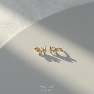 May include: A pair of gold-colored leaf-shaped stud earrings with clear, sparkling accents. The earrings are displayed on a white surface, with the brand name "Winnies & ATOWN - JEWELRY" visible at the bottom.