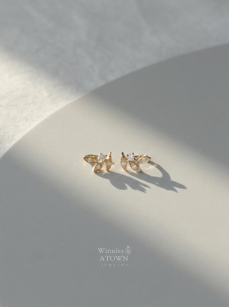 May include: A pair of gold-colored leaf-shaped stud earrings with clear, sparkling accents. The earrings are displayed on a white surface, with the brand name "Winnies & ATOWN - JEWELRY" visible at the bottom.