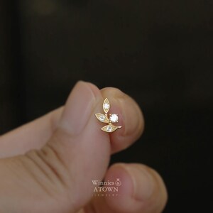 May include: A gold-colored earring with a leaf design, featuring small clear gemstones. The earring has a post for pierced ears. The brand name "Winnies & ATOWN - jewelry" is visible.