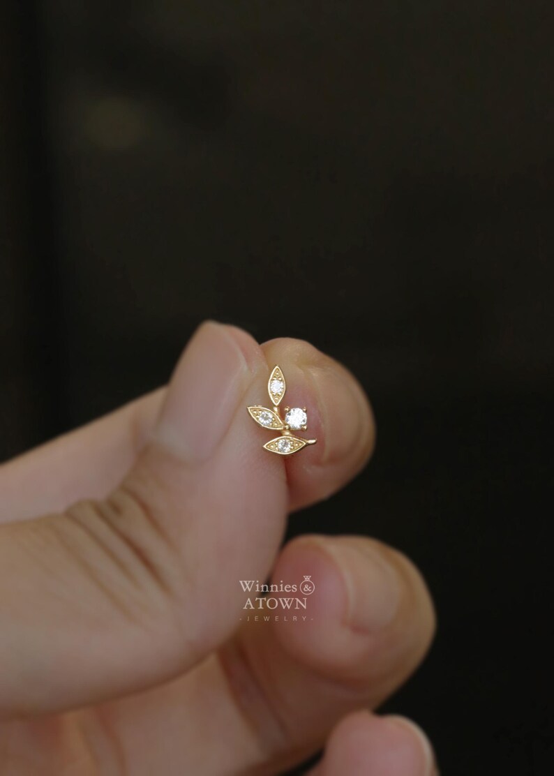 May include: A gold-colored earring with a leaf design, featuring small clear gemstones. The earring has a post for pierced ears. The brand name "Winnies & ATOWN - jewelry" is visible.
