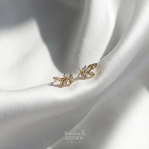 May include: A pair of gold-colored leaf-shaped stud earrings with small, clear gemstones. The earrings are displayed on a white, silky fabric. The brand name "Winnies & ATOWN JEWELRY" is visible in the lower right corner.
