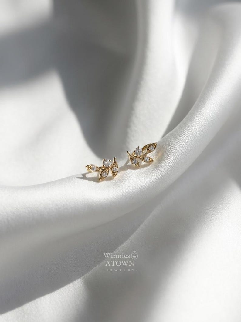May include: A pair of gold-colored leaf-shaped stud earrings with small, clear gemstones. The earrings are displayed on a white, silky fabric. The brand name "Winnies & ATOWN JEWELRY" is visible in the lower right corner.