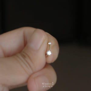 Puede incluir: Un peque&ntilde;o piercing para el ombligo de color plateado con dos piedras preciosas redondas y transparentes. La joya se sostiene entre los dedos sobre un fondo borroso. La marca "Winnies ATOWN JEWELRY" es visible.