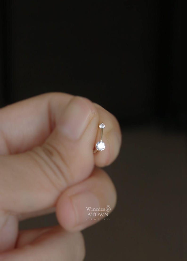 Puede incluir: Un peque&ntilde;o piercing para el ombligo de color plateado con dos piedras preciosas redondas y transparentes. La joya se sostiene entre los dedos sobre un fondo borroso. La marca "Winnies ATOWN JEWELRY" es visible.