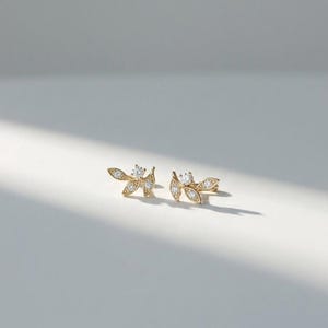 May include: A pair of gold-colored leaf-shaped earrings with small, sparkling clear gemstones. The earrings are displayed on a white surface with soft lighting. The earrings have a delicate, nature-inspired design.