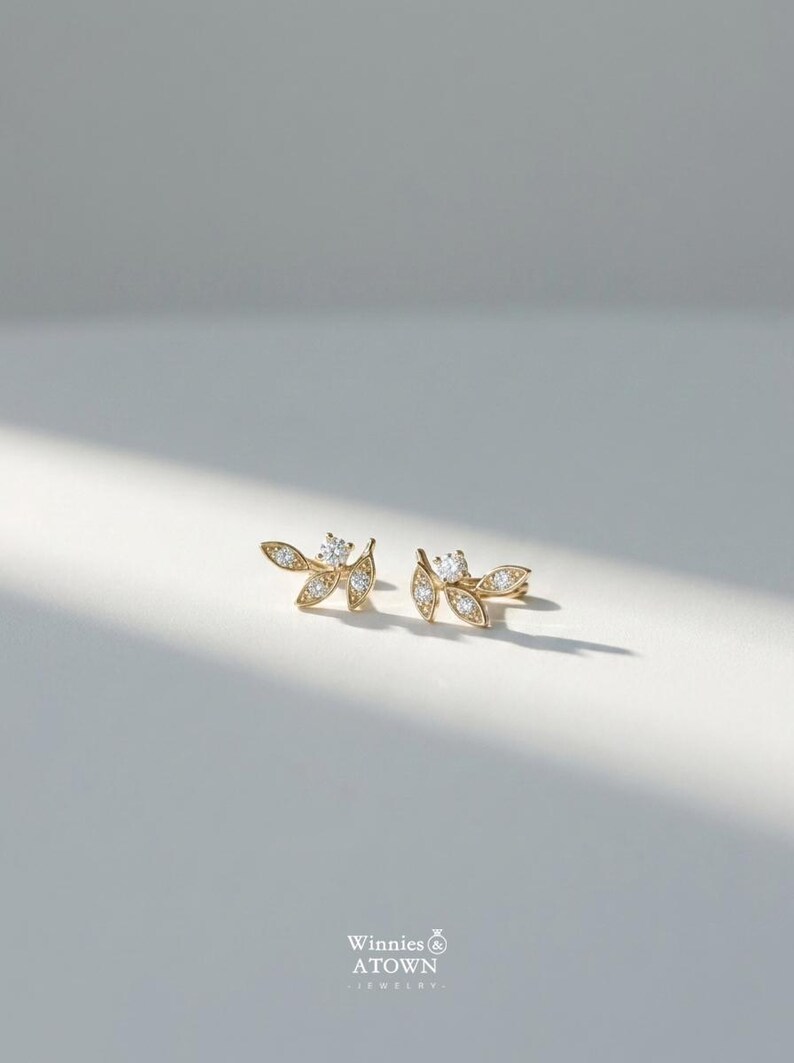May include: A pair of gold-colored leaf-shaped earrings with small, sparkling clear gemstones. The earrings are displayed on a white surface with soft lighting. The earrings have a delicate, nature-inspired design.