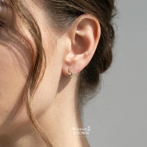 May include: A close-up of a person's ear adorned with a delicate gold stud earring. The earring features a leaf-like design with small, sparkling gemstones. The jewelry is from Winnies & ATOWN JEWELRY.