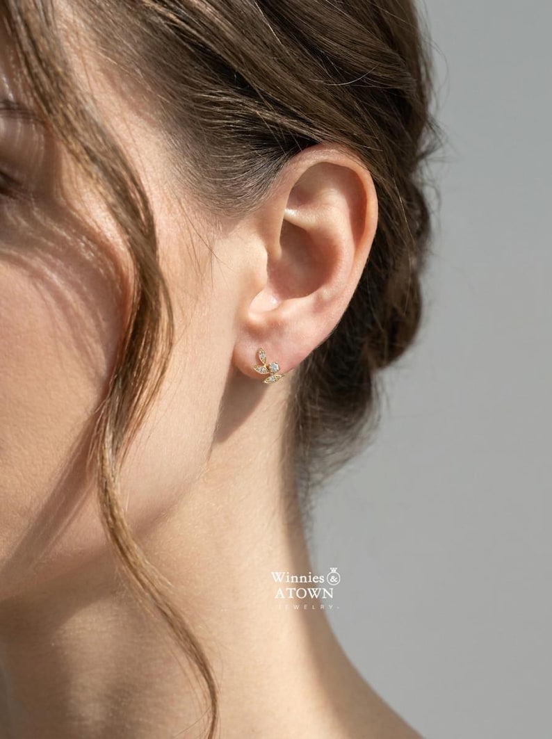 May include: A close-up of a person's ear adorned with a delicate gold stud earring. The earring features a leaf-like design with small, sparkling gemstones. The jewelry is from Winnies & ATOWN JEWELRY.