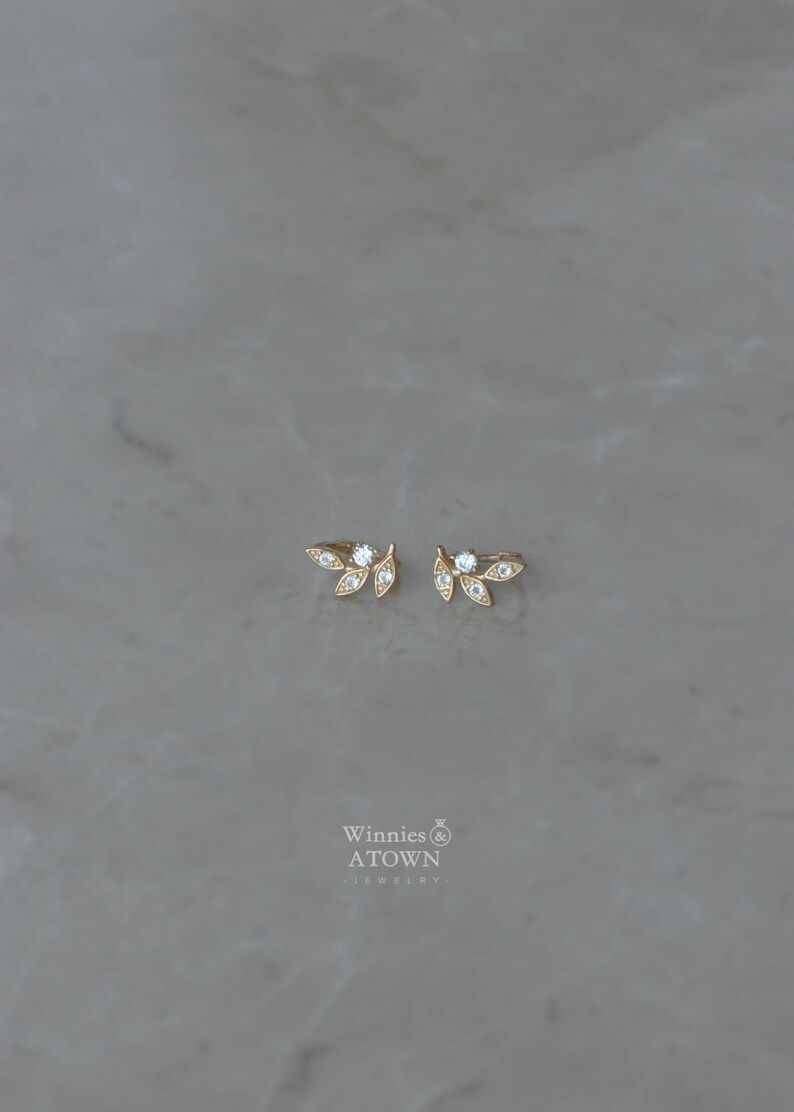 May include: A pair of gold-colored leaf-shaped earrings with small, clear gemstones. The earrings are displayed on a gray surface. The brand name "Winnies & ATOWN - jewelry" is visible in the lower center.