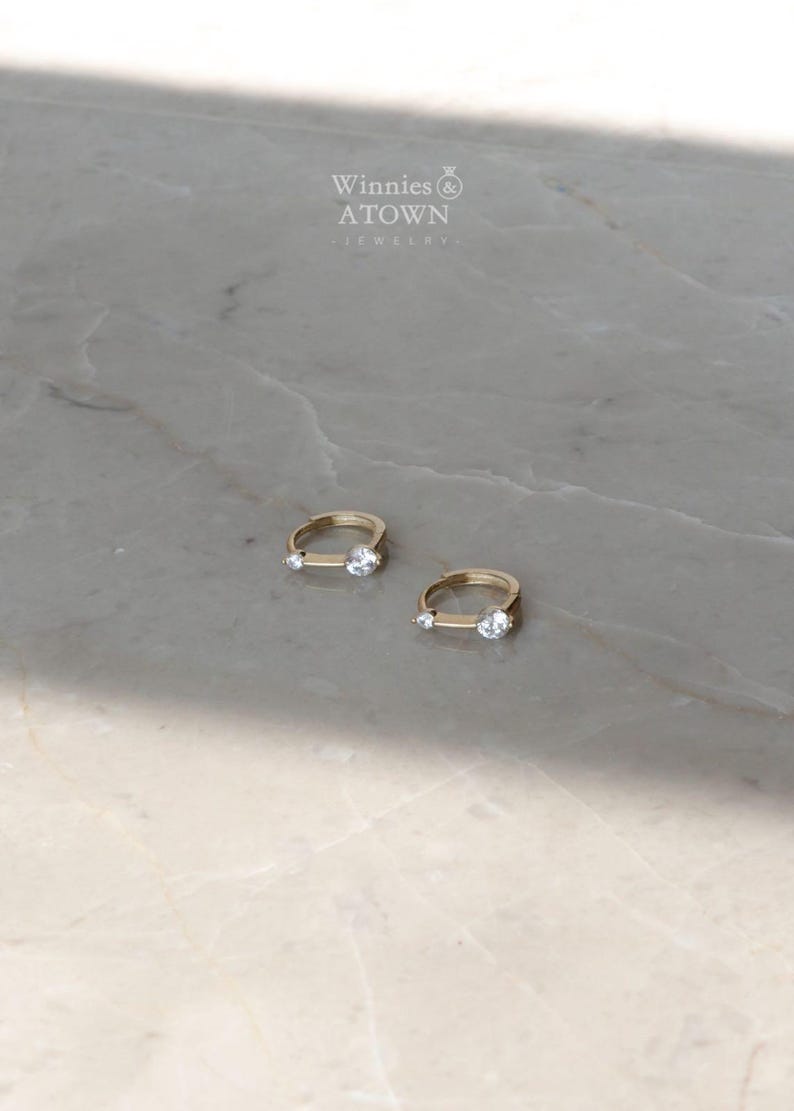 Puede incluir: Dos pendientes de aro dorados, cada uno con una peque&ntilde;a gema transparente y una gema transparente y redonda m&aacute;s grande. Los pendientes se muestran sobre una superficie clara, similar al m&aacute;rmol. La marca "Winnies & ATOWN JEWELRY" es visible en el fondo.