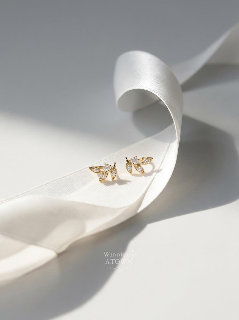 May include: A pair of gold-colored leaf-shaped stud earrings with small, clear gemstones. The earrings are displayed on a white ribbon against a white background. The brand name "Winnies ATown" is visible.