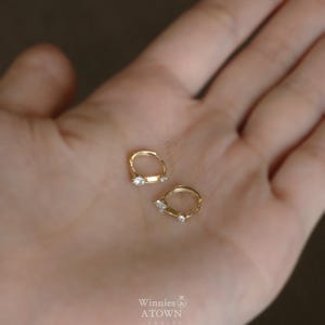 Puede incluir: Un par de pendientes de aro peque&ntilde;os, de color dorado, cada uno con una sola gema transparente. Los pendientes se muestran sobre un fondo claro. La marca "Winnies ATOWN" es visible en la esquina inferior derecha.