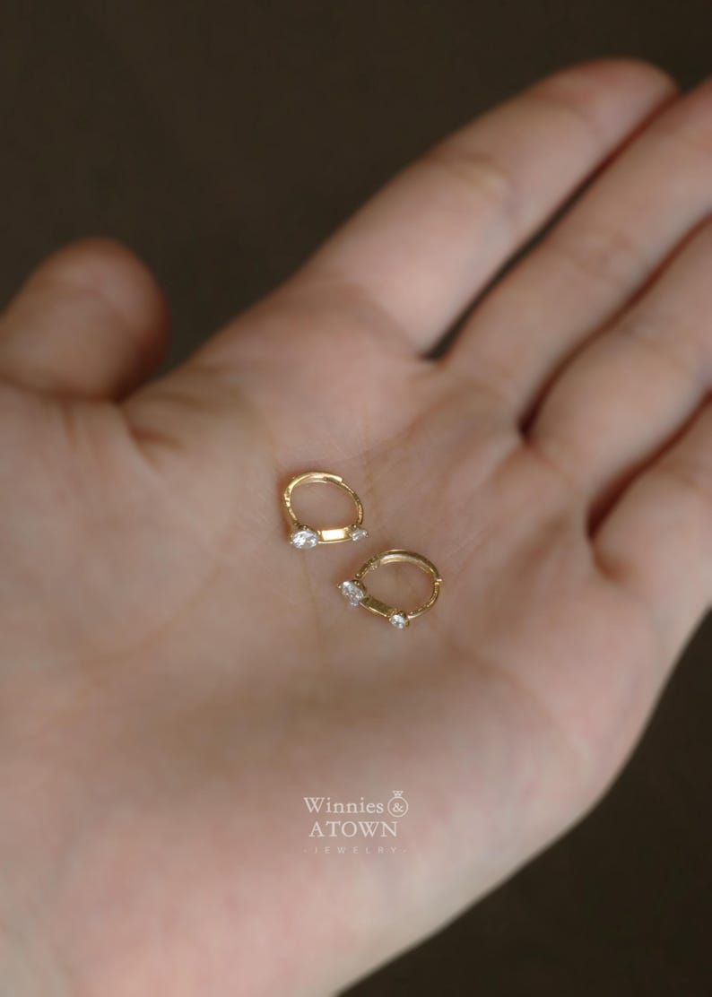 Puede incluir: Un par de pendientes de aro peque&ntilde;os, de color dorado, cada uno con una sola gema transparente. Los pendientes se muestran sobre un fondo claro. La marca "Winnies ATOWN" es visible en la esquina inferior derecha.