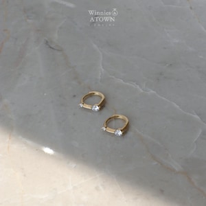Puede incluir: Dos pendientes de aro dorados, cada uno con una peque&ntilde;a gema transparente. Se exhiben sobre una superficie de m&aacute;rmol gris claro y blanco. La marca "Winnies & ATOWN JEWELRY" es visible en el fondo.