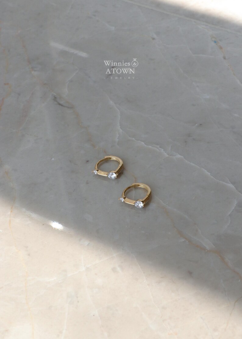 Puede incluir: Dos pendientes de aro dorados, cada uno con una peque&ntilde;a gema transparente. Se exhiben sobre una superficie de m&aacute;rmol gris claro y blanco. La marca "Winnies & ATOWN JEWELRY" es visible en el fondo.